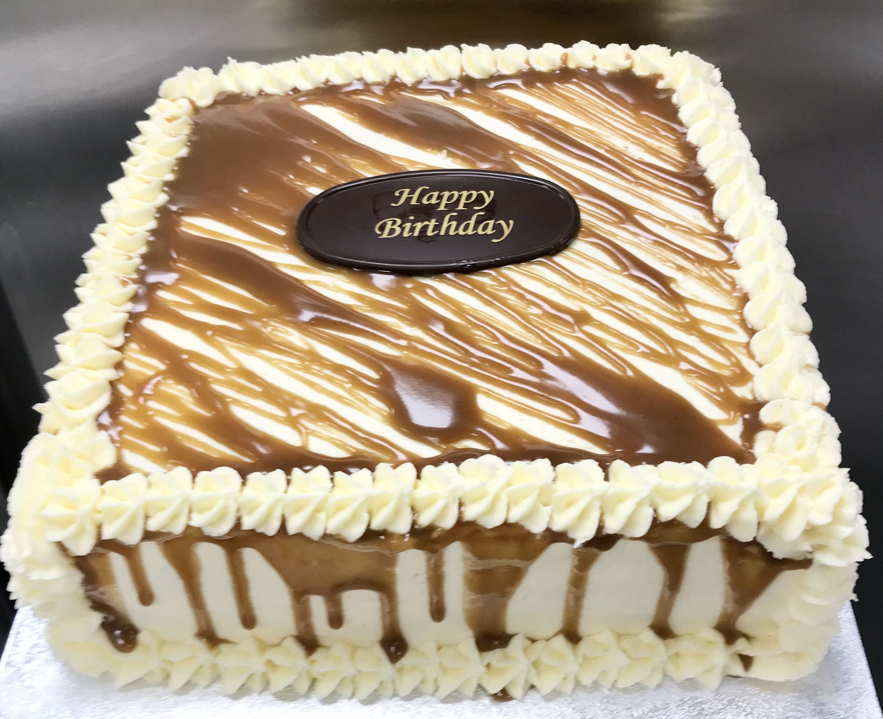 A square birthday cake topped with homemade salted caramel sauce, bordered with white frosting swirls. A chocolate plaque in the center reads 'Happy Birthday.' The sides of the Salted Caramel Cake are decorated with vertical drips of caramel alternating with white frosting.